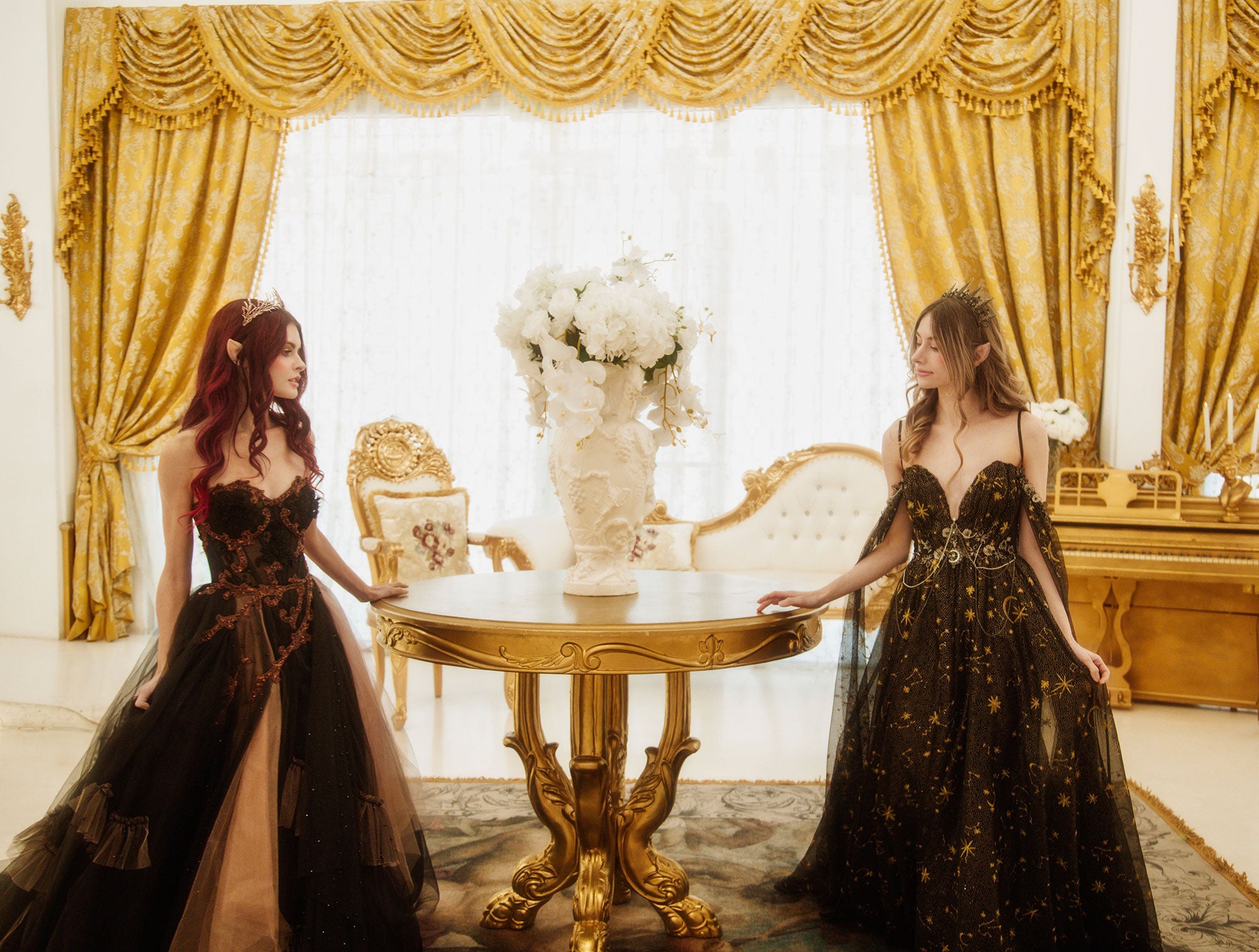 Two fae princesses in ornate black celestial themed gowns walk around a table in a golden palace and talk about The Paravelle Ball fantasy event.